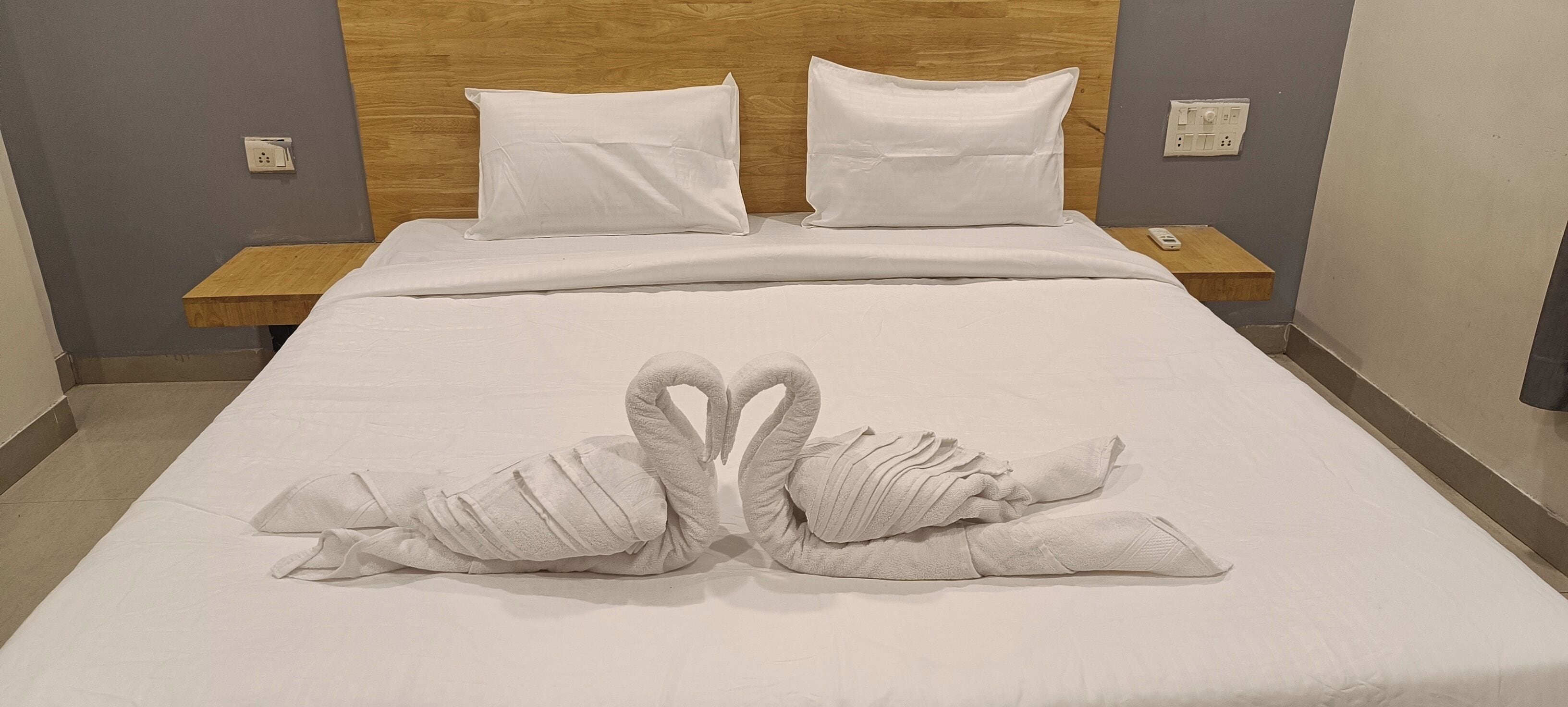 Un lit bien fait, agrémenté d’une décoration en forme de deux cygnes réalisée avec des serviettes, une tête de lit en bois et des tables de chevet avec des lampes.