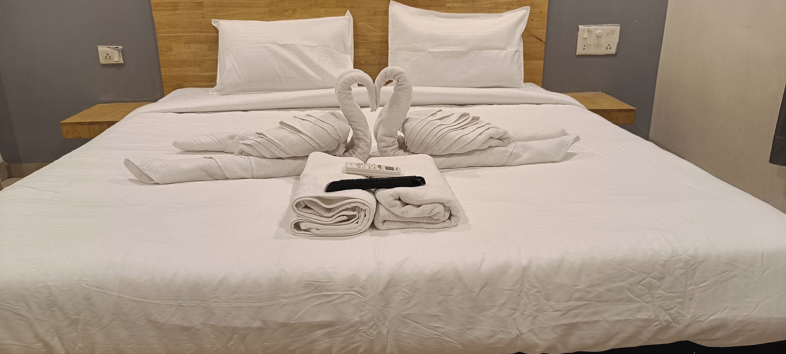 Una cama bien hecha con toallas dispuestas en forma de corazón, un termómetro digital y un objeto negro sobre la cama.