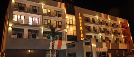 Un hôtel à plusieurs étages, doté d’une façade moderne, de fenêtres illuminées et d’une enseigne bien visible portant le nom « IMPERIUM ».