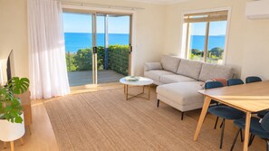A living room with a sofa, a coffee table, and a dining table with chairs. There is a large sliding glass door leading to a balcony with a view of the sea.
