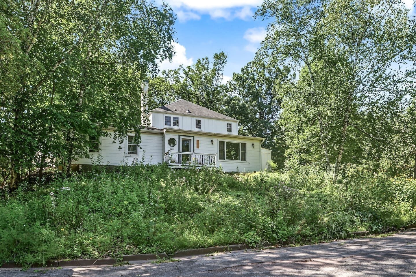 A two-story house with a porch, surrounded by dense greenery.