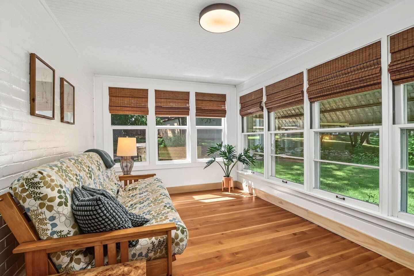 A room with a wooden floor, a sofa with patterned cushions, a lamp, and large windows with bamboo shades.