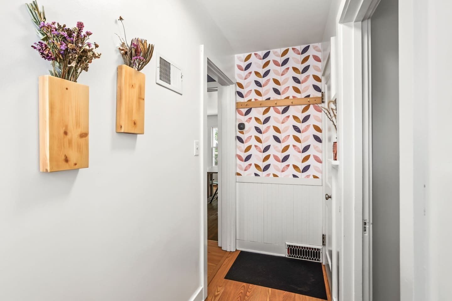 Un pasillo con una puerta de madera, un felpudo negro en la entrada y dos macetas de madera fijadas a la pared con flores secas.
