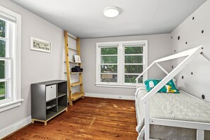 A child's bedroom with a bunk bed, a wooden ladder, a desk, and a window with a view of greenery.