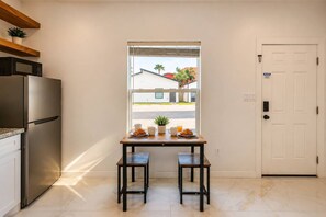 A kitchen with a dining table, two chairs, a refrigerator, and a window with a view of buildings and palm trees. - #14 | Island Studio Escape | Pool+Wi-Fi | Beach (South Padre Island)