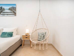 Ein Schlafzimmer mit einem Bett, einem Nachttisch mit Lampe, einem Hängekorbstuhl aus Makramee und einem Bild an der Wand.