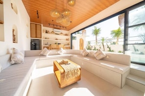 Uma sala de estar moderna com um sofá grande, uma mesa de centro de madeira e uma área de cozinha com geladeira.