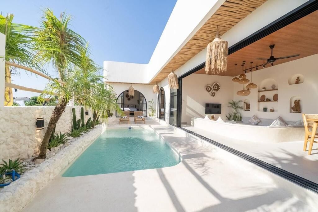 A modern outdoor pool area with a stone wall, palm trees, and a seating area with chairs and a table.