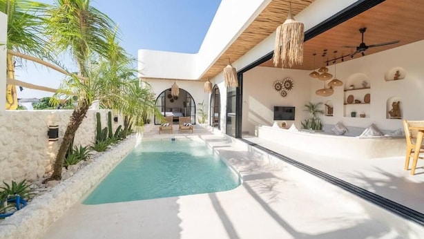 A modern outdoor pool area with a stone wall, palm trees, and a seating area with chairs and a table. - Disenta Villa (Badung)