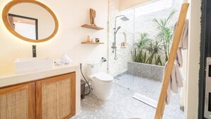 A modern bathroom with a freestanding toilet, a sink with a mirror, and a shower area with a glass door. - Disenta Villa (Badung)