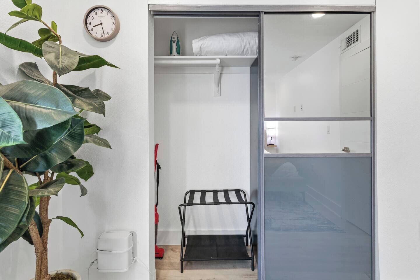 A compact room with a bed, a small shelf with a white pillow, a clock on the wall, and a potted plant.