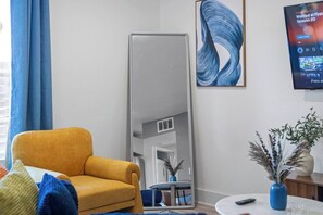 Ein Wohnzimmer mit einem gelben Sessel, einem großen Spiegel, einem blauen abstrakten Gemälde und einem Fernseher, der eine Sendung zeigt.