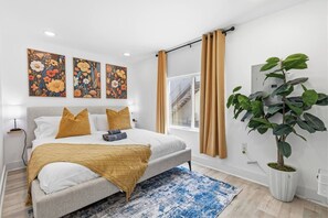 Ein modernes Schlafzimmer mit Bett, großem Fenster mit Vorhängen, einer Topfpflanze und Bildern an der Wand.