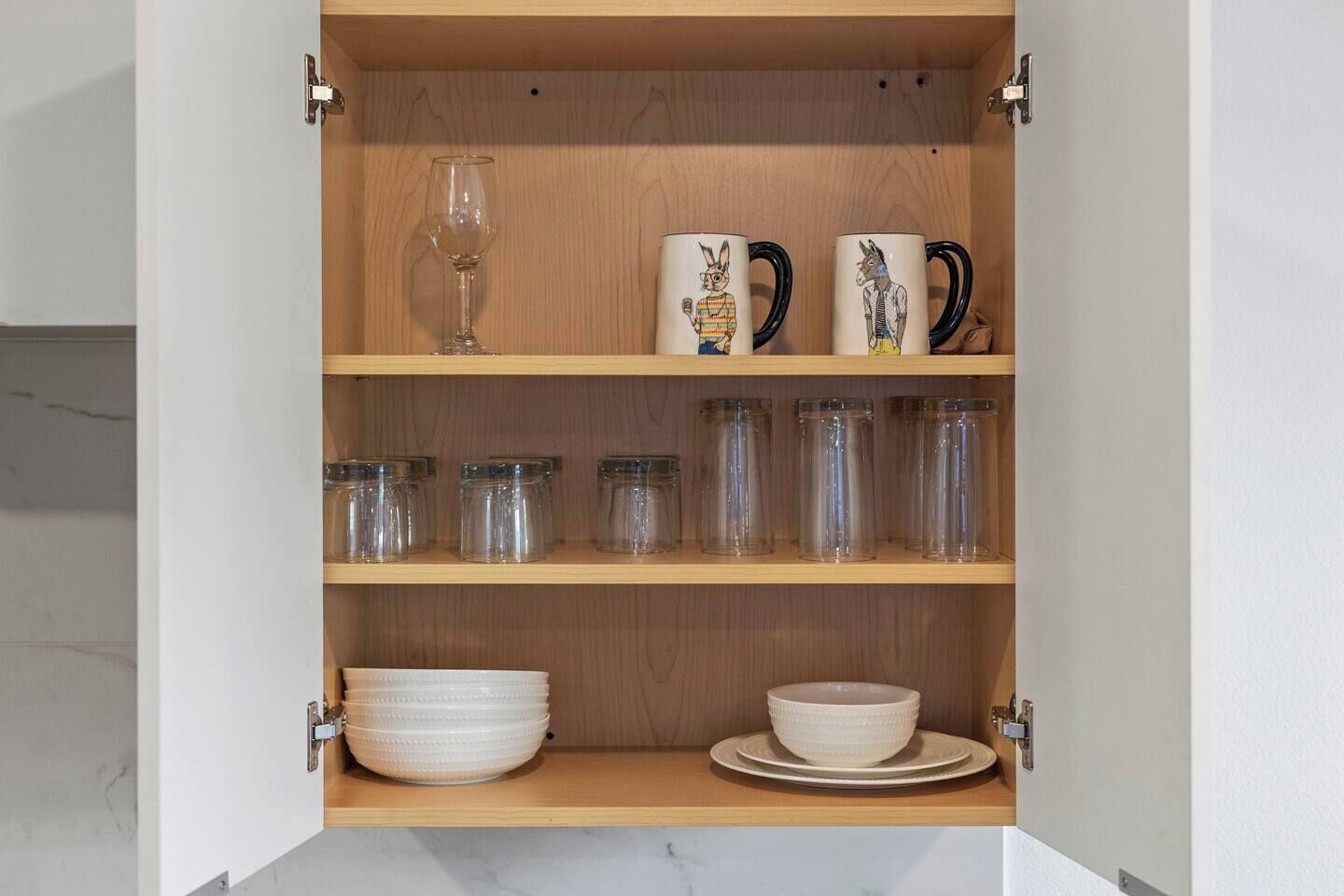 A kitchen cabinet with shelves containing glassware, mugs, and storage jars.