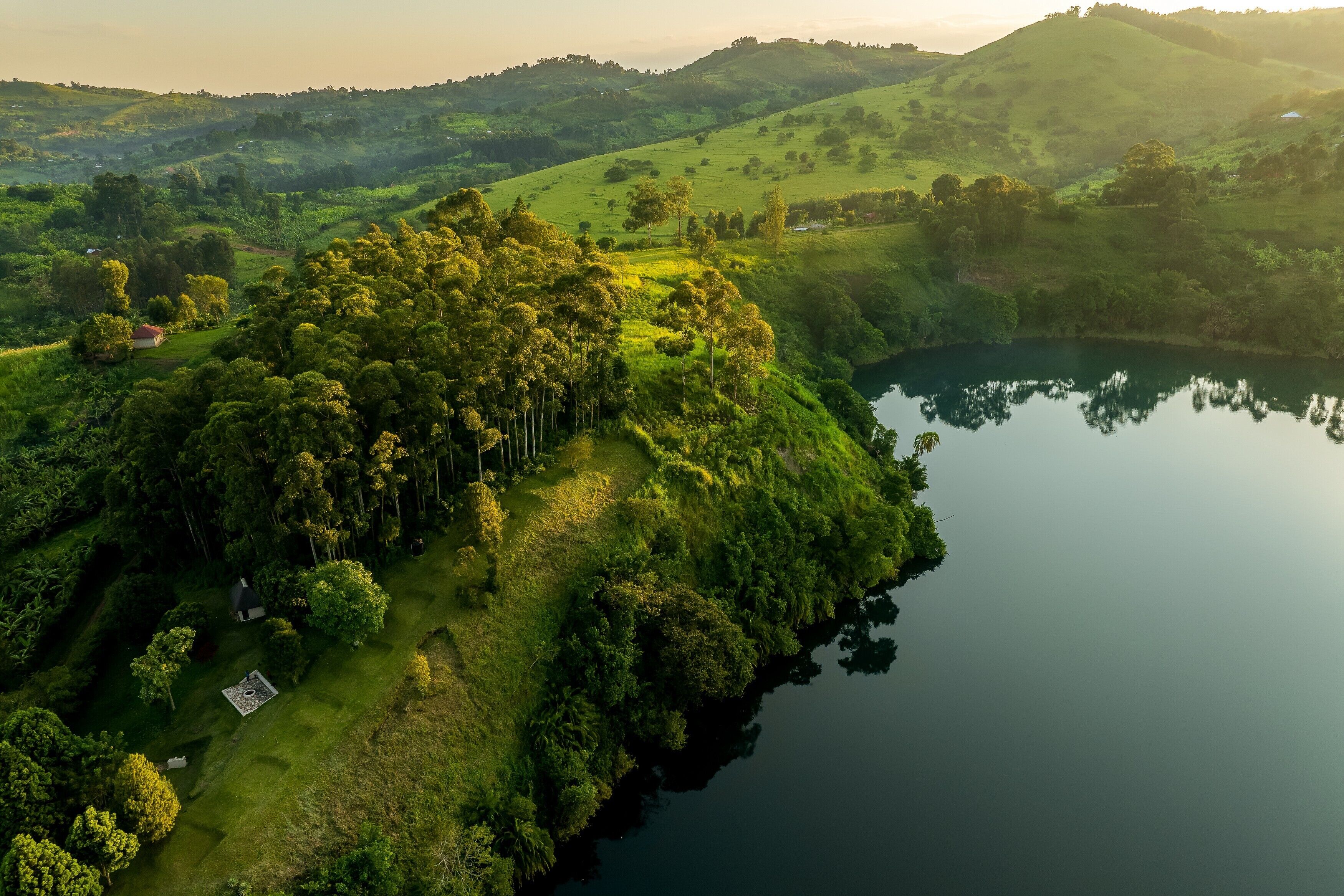 Vista aérea de un paisaje verde y exuberante con un lago, colinas y un pequeño edificio con techo rojo.