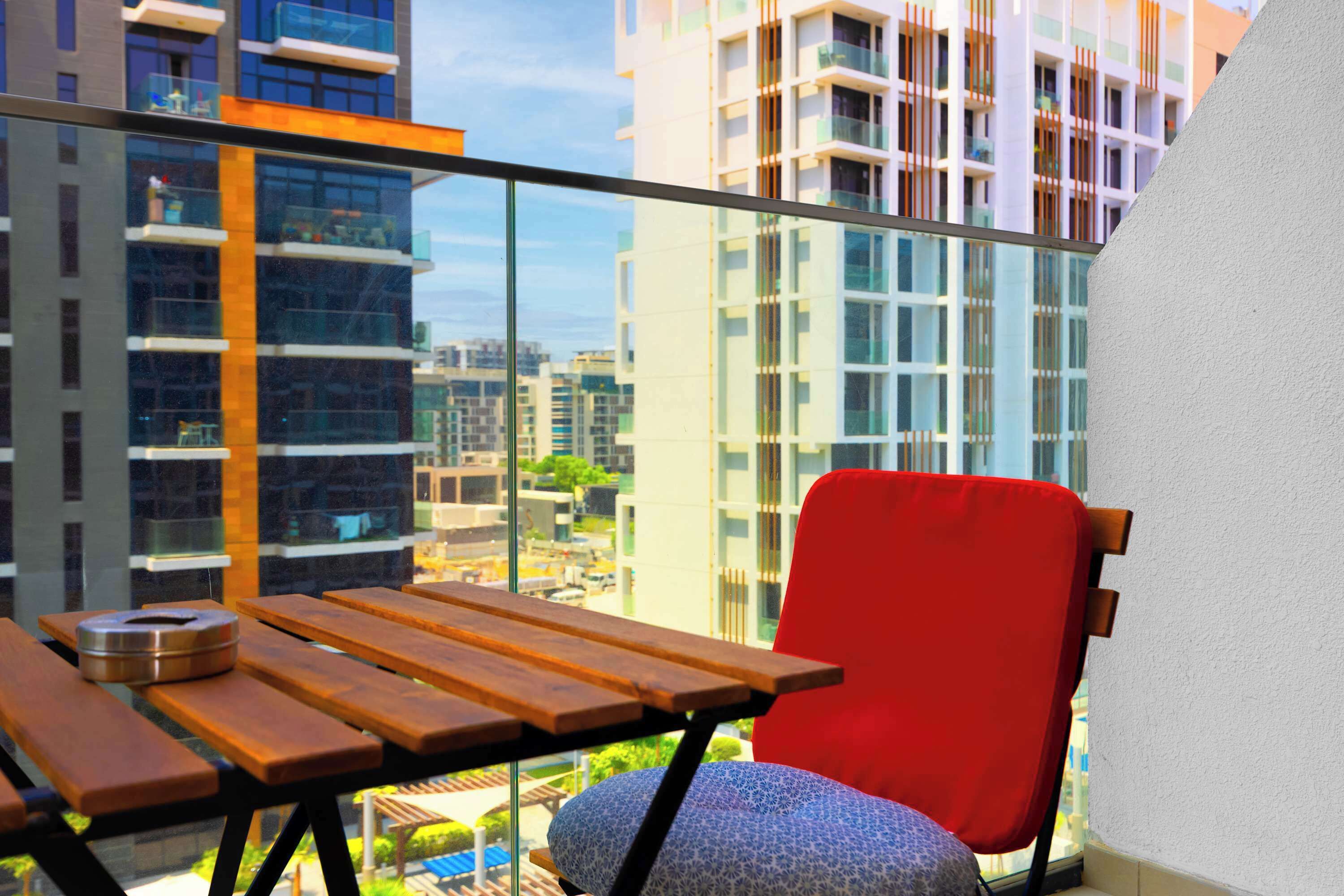 Een balkon met een houten tafel en een rode stoel, met uitzicht op een stadsbeeld met moderne gebouwen.