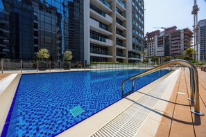 A swimming pool with a tiled edge, surrounded by a wooden deck and modern buildings. - Riviera 42 Studio · Lagoon Pool · Meydan (Dubai)
