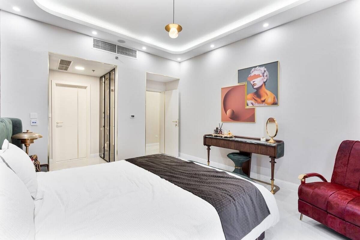 A modern hotel room with a large bed, a vanity with a mirror, a red armchair, and artwork on the wall.