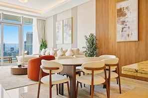 A modern living room with a large sofa, dining table, and chairs, large windows with a city view, and decorative plants. - Imperial 3BR · Canal View · Downtown (Dubai)