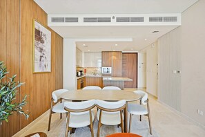 A modern dining area with a round table and six chairs, a kitchen in the background, and a potted plant on the left. - Imperial 3BR · Canal View · Downtown (Dubai)