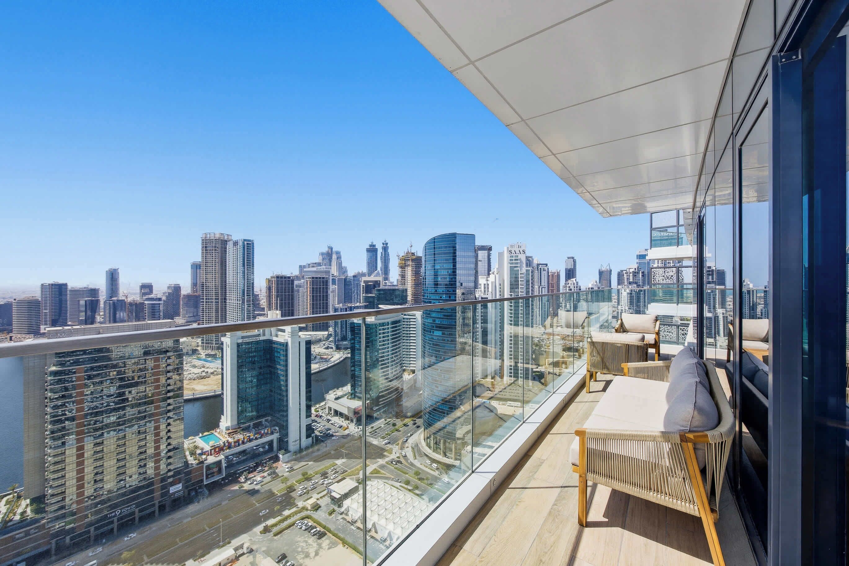 Un balcon situé dans un immeuble de grande hauteur, offrant une vue sur la ville, aménagé avec un canapé et des fauteuils, et doté d’une rambarde en verre.