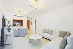 A modern living room with a sofa, a coffee table, and a TV. There is a plant and a dining area with chairs. - Blvd Point 2BR · Burj View · Dubai Mall (Dubai)