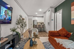 A modern living room with a flat-screen TV, a sofa, a coffee table, and a dining area with chairs and a kitchen in the background. - Near NRG Stadium & DT| Pool & Gym, Free Parking (Houston)