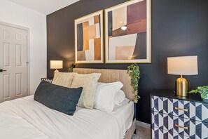 A bedroom with a bed, two bedside lamps, a decorative wall piece, and a geometric-patterned cabinet. - Near NRG Stadium & DT| Pool & Gym, Free Parking (Houston)