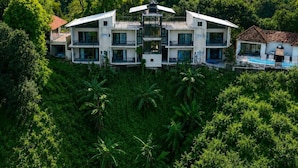 Un edificio moderno de varios pisos con balcones, rodeado de abundante vegetación y un cielo despejado.