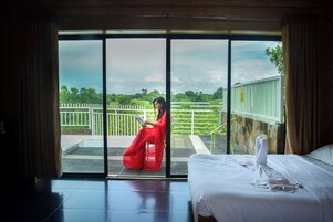 A hotel room with a large sliding glass door leading to a balcony with a view of a green landscape and a white fence.