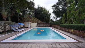 Una piscina rectangular con borde de azulejos, rodeada por un muro de piedra y un seto bajo. Hay sillas de descanso y sombrillas a lo largo de la piscina.