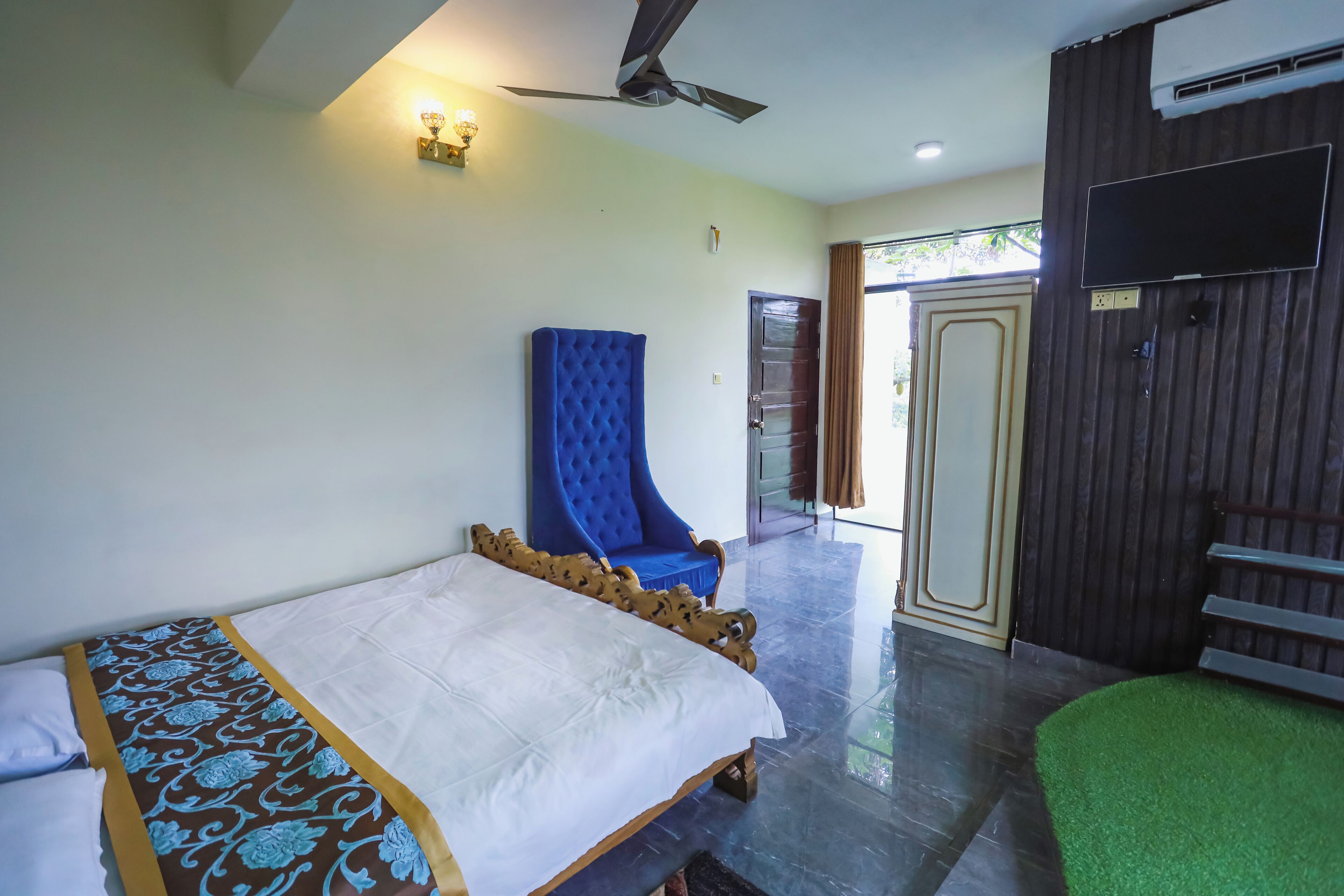 A hotel room with a bed, a blue armchair, a ceiling fan, and a TV mounted on the wall.