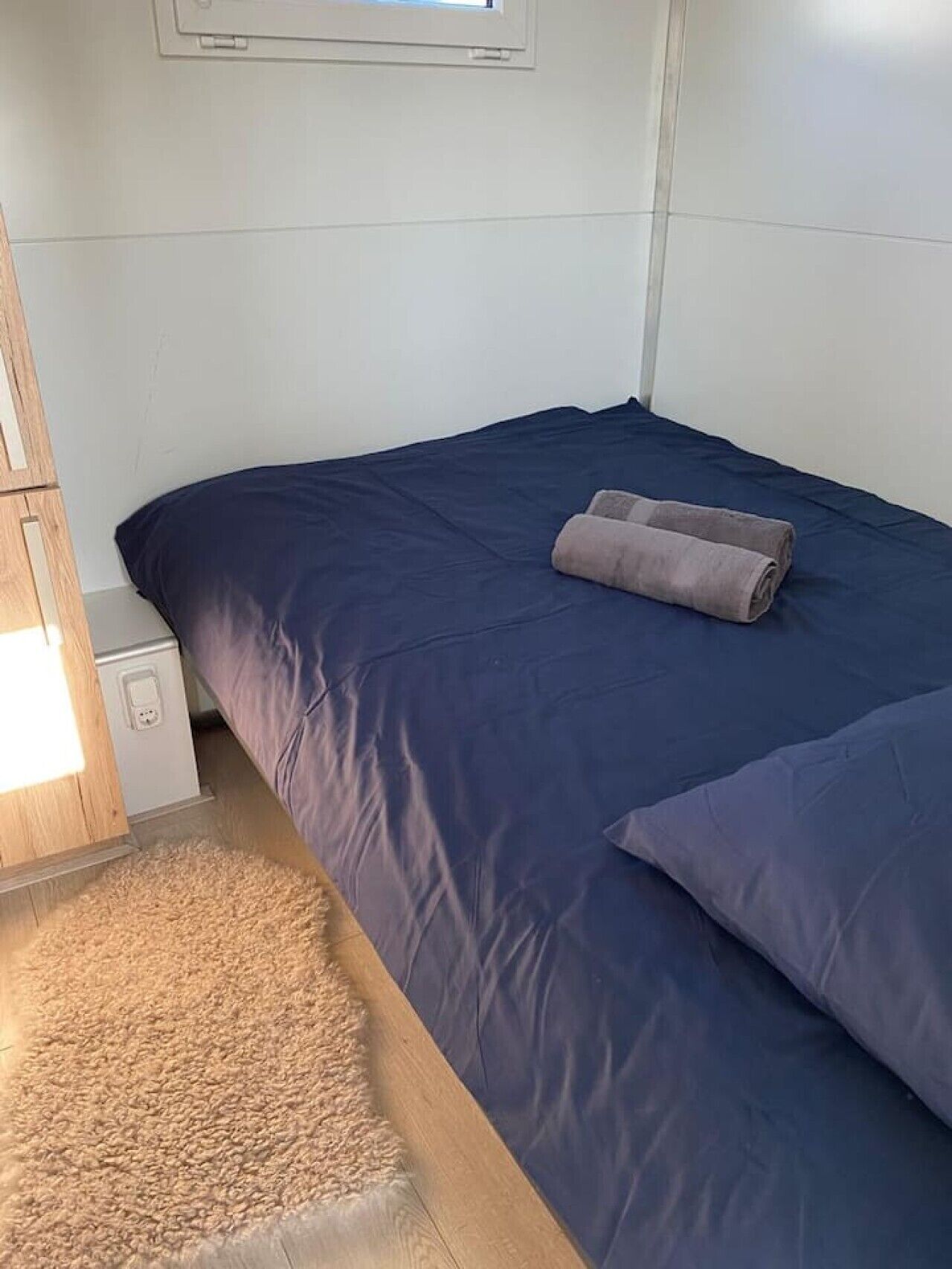 Een klein, net bed met een blauwe sprei en twee kussens, een houten nachtkastje met een lamp, en een zacht vloerkleed.