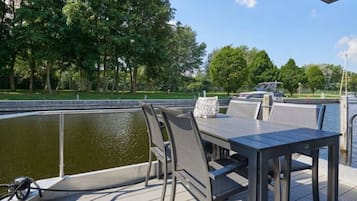 Una terraza exterior con mesa y sillas, con vista a un cuerpo de agua y un parque con árboles.
