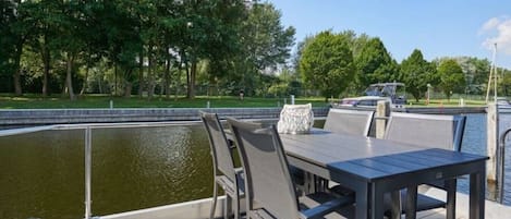 En udendørs terrasse med et bord og stole, med udsigt over vand og en park med træer.
