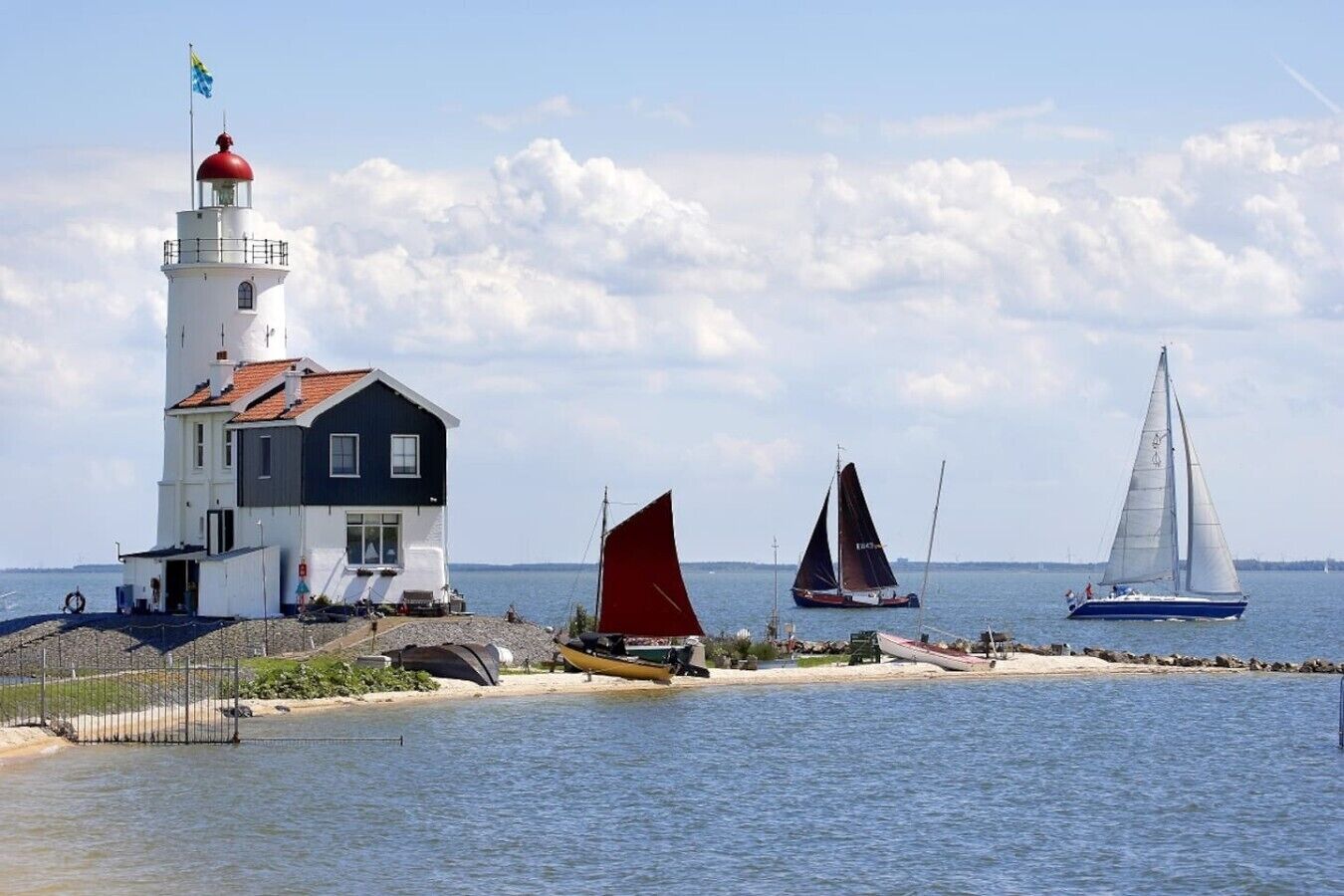 Ein Leuchtturm mit roter Spitze, ein weißes Gebäude mit dunklem Dach und Segelboote auf dem Wasser.