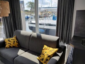 Un canapé gris avec deux coussins jaunes, une fenêtre donnant sur des bateaux et des bâtiments, et un meuble sombre.