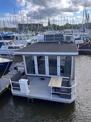 Ein schwimmendes Hausboot liegt in einer Marina festgemacht, mit Deck, Sitzgelegenheit und Tisch.