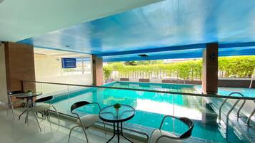 An indoor swimming pool with clear glass walls, surrounded by chairs and tables, and a view of greenery outside.