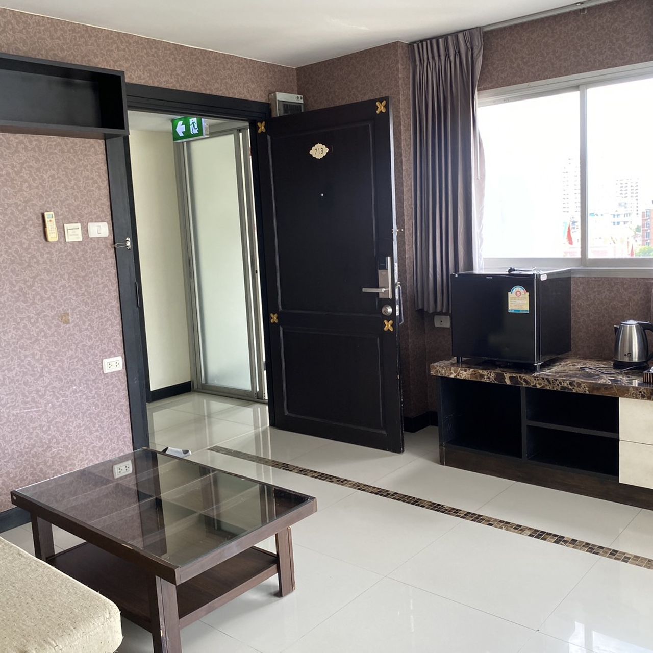 A hotel room with a glass-top table, a dark door with a number, a kitchenette with a coffee maker, and a window with a view of buildings.