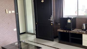 A hotel room with a glass-top table, a dark door with a number, a kitchenette with a coffee maker, and a window with a view of buildings.