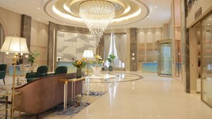 A hotel lobby with a reception desk, green chairs, a chandelier, and a glass elevator.