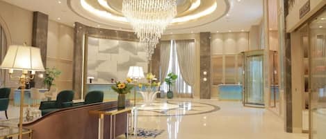 A hotel lobby with a reception desk, green chairs, a chandelier, and a glass elevator.