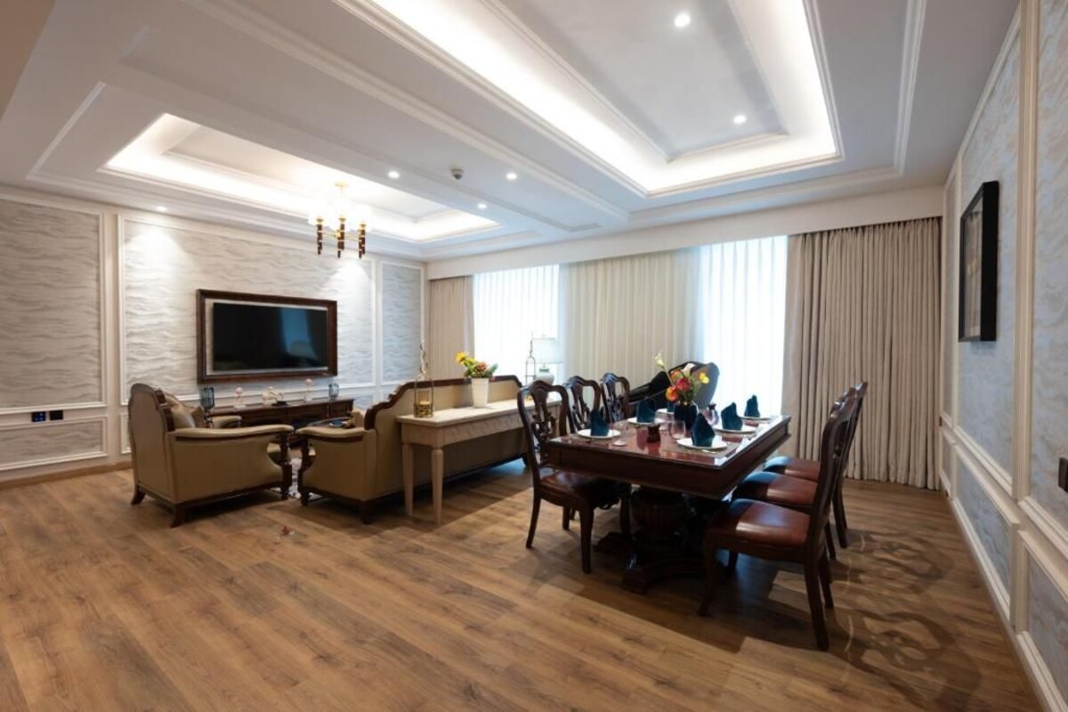 A spacious living room with a modern chandelier, a flat-screen TV mounted on the wall, a dining area with a wooden table and chairs, and a large window with curtains.