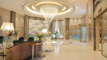 A hotel lobby with a reception desk, green chairs, a chandelier, and a glass elevator.