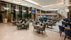 A spacious hotel lobby with multiple dining areas, a reception desk, and a staircase leading to an upper level.
