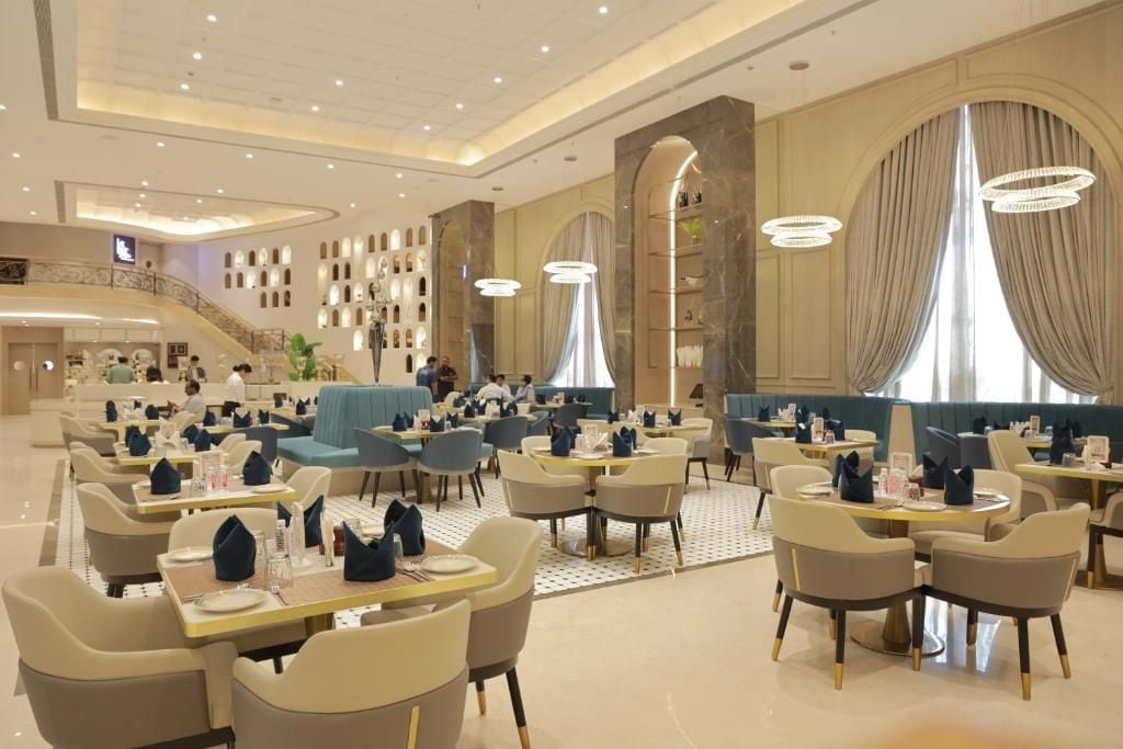 Un amplio comedor de hotel con varias mesas y sillas, una escalera central e iluminación decorativa.