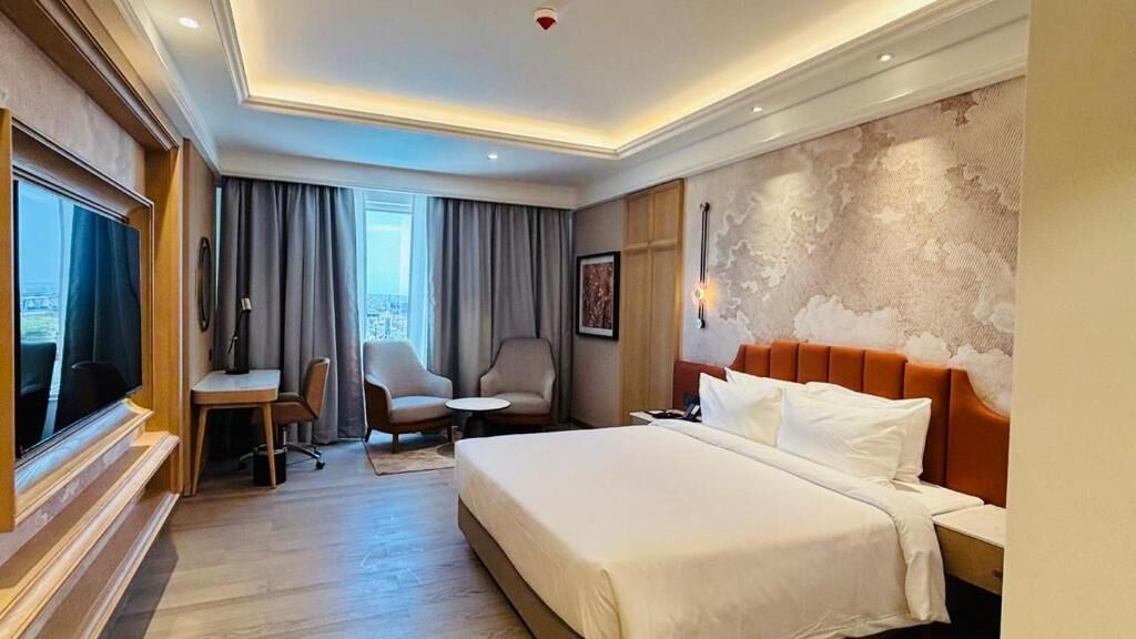 A hotel room with a large bed, a desk, a chair, a small table, and a television.