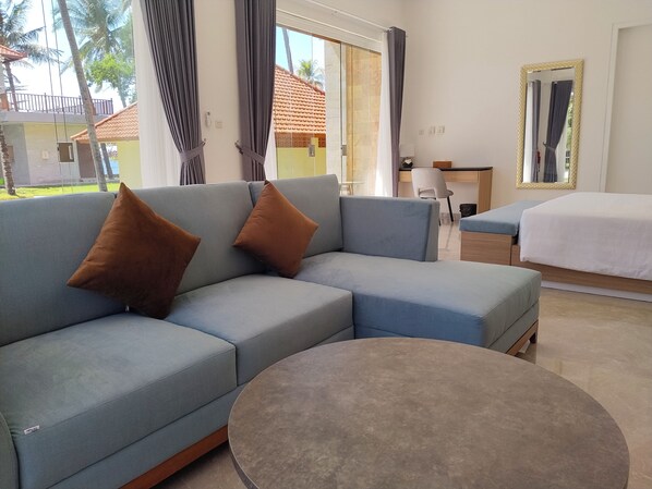 A modern hotel room with a sectional sofa, a round coffee table, a bed, and a desk with a chair.