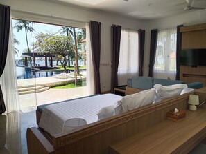 A hotel room with a bed, a sofa, a TV, and a view of a pool and palm trees. - Scampi Indorus Resort (Tejakula)